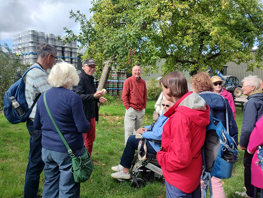 Group of people gathered outdoors at Sheppy’s Cider Farm, listening to a guide beside stacks of cider kegs.